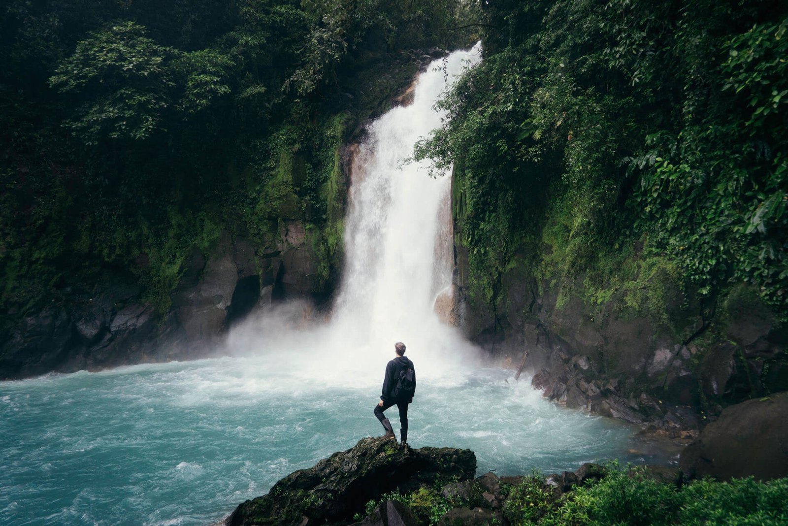 Person with a backpack stands on a rocky ledge watching a tall waterfall surrounded by dense jungle.