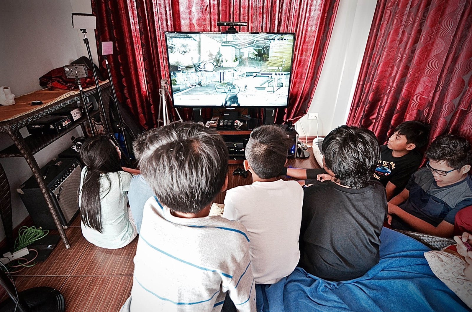 Group of children sitting on the floor, facing a large TV screen playing a video game in a cluttered living room setup with red curtains behind.