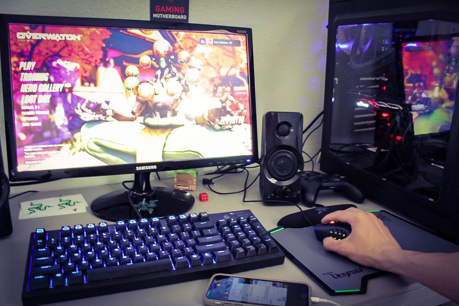 Gaming desk setup with a colorful game on a Samsung monitor, blue RGB keyboard, and a wired mouse under a clear side-window PC case.