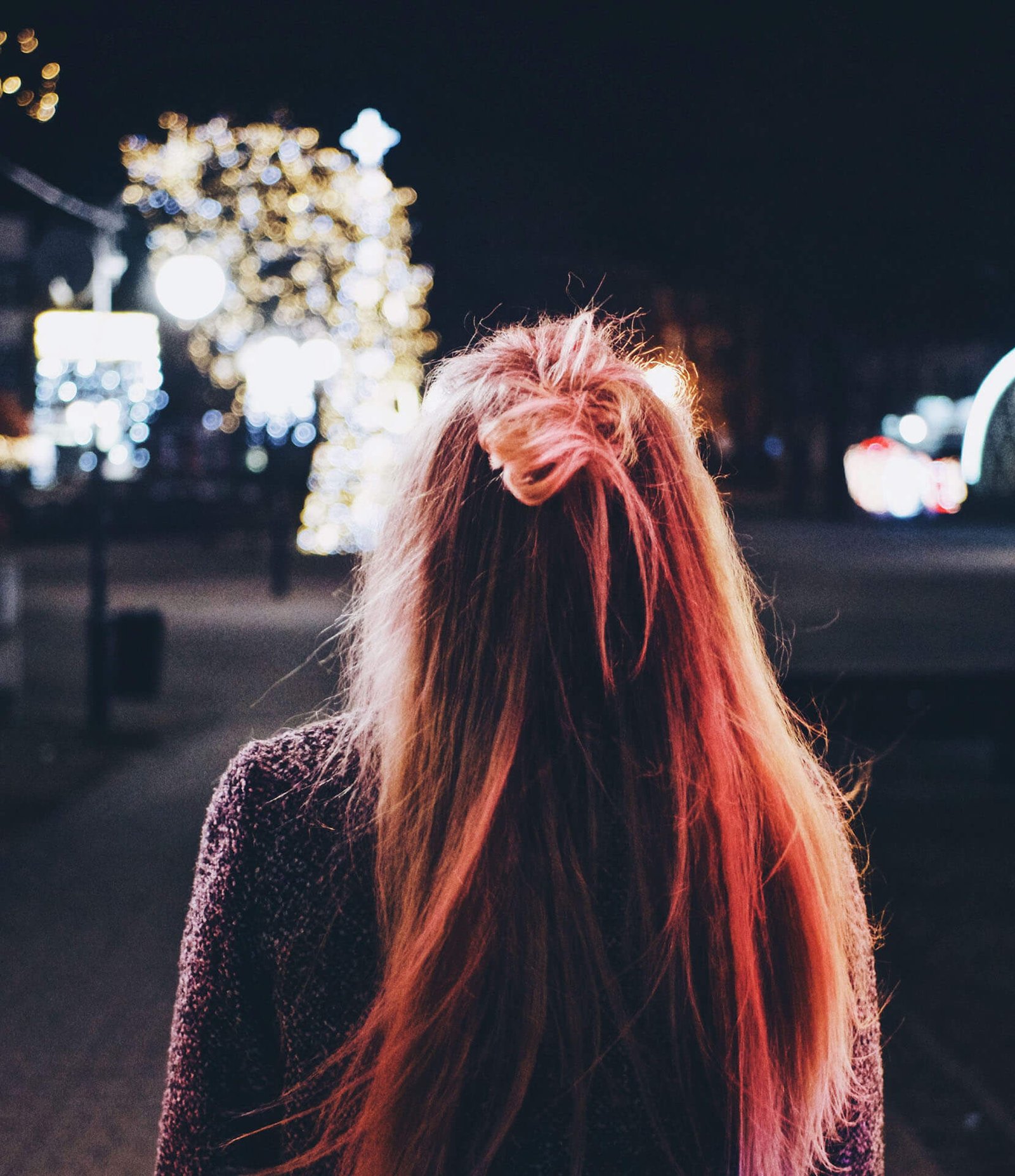 Back view of a person with pink hair standing in a nighttime city scene with festive, blurred lights in the background