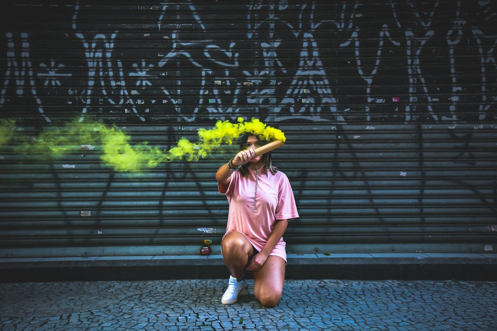 Woman in a pink dress kneels on a cobblestone street, holding a yellow smoke flare in front of a graffiti-covered metal shutter.