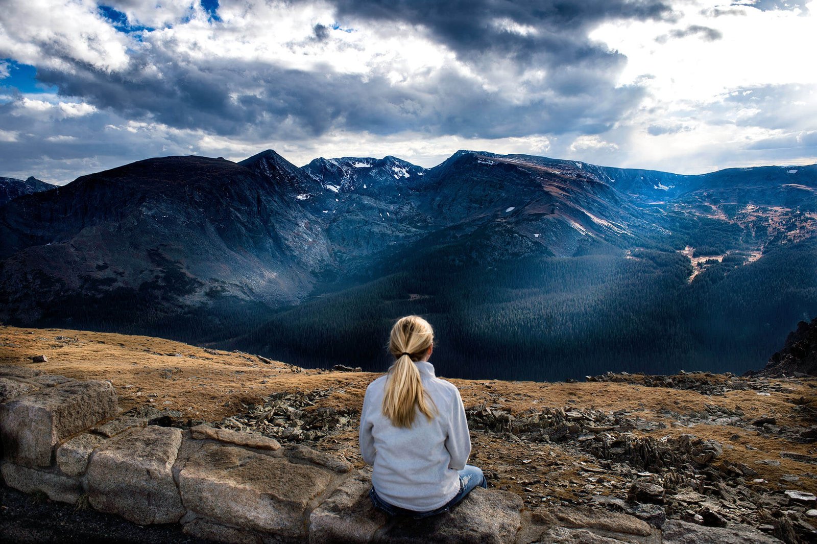 Person with blonde hair sits on rocky ledge overlooking a mountain range under a cloudy sky.